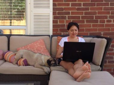 pets | Neuromyelitis News | Jennifer sits on a couch in her backyard working on her laptop. Her fawn pug lies next to her, his leg kicked over a pillow.