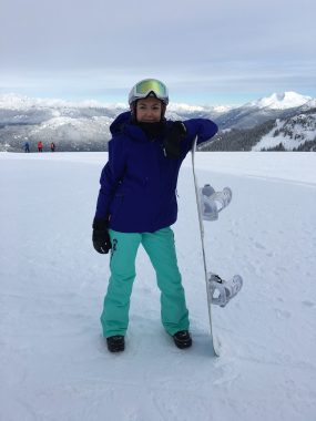 snowboarding with a disability | Neuromyelitis News | Jennifer wears blue and green snow gear and a reflective visor and leans against her snowboard while standing atop the snowy peak of Whistler Mountain.