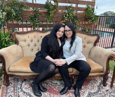 friendships with NMO | Neuromyelitis News | Jennifer's friend Chan poses with her wife, Jessica, on a couch placed outdoors in front of a wall of plants.