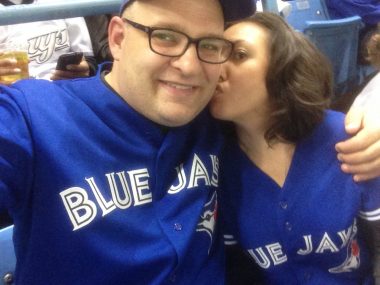 COVID-19 Restrictions | Neuromyelitis News | Jennifer gives her husband, Mike, a kiss on the cheek during a Toronto Blue Jays home opener in 2015. Both are wearing Blue Jays jerseys.