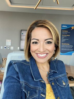 find dream jobs | Neuromyelitis News | photo of Candice, seated, wearing a denim jacket with a yellow shirt. A DaVita poster is behind her.