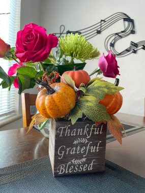 A holiday centerpiece with small pumpkins and roses, and the words "Thankful, Grateful, Blessed" sits on a dining room table 