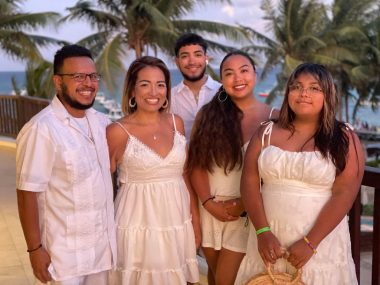 A family of five — two men and three women — poses on a beach with palm trees in the background during a summer family vacation. All are wearing white - the women white dresses and the men white guayabera shirts. The scenery, clothing, and smiles make for a stunning and warm family photo.