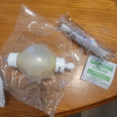 Photo of an Eclipse portable medication infusion pump, a saline tube, and an alcohol wipe. The pump, which looks like a small, white balloon with white plastic caps on each end, is packaged in a small, clear plastic bag. The saline tubes are also bunched up in a plastic bag, and the alcohol swabs are just in a Band-Aid-like wrapping. 