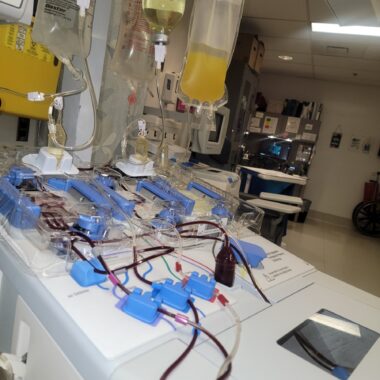 On a table, devices in what seem to be pastel blue and clear plastic have tubes running from them, most seemingly carrying blood. An IV bag with yellow fluid is above the table.