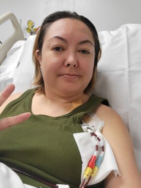 A woman in a forest-green tank top is sitting at an angle in a hospital bed. Two tubes seemingly carrying blood are coming from beneath her shirt; they have brightly colored connectors.