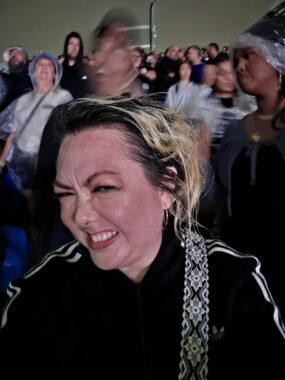 A woman squints and smiles for a selfie, surrounded by a crowd of people at a large outdoor venue.