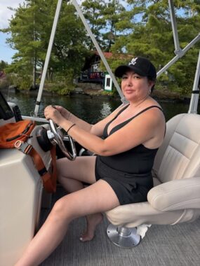A woman wearing a black swimsuit and baseball cap sits behind the wheel of a powerboat.